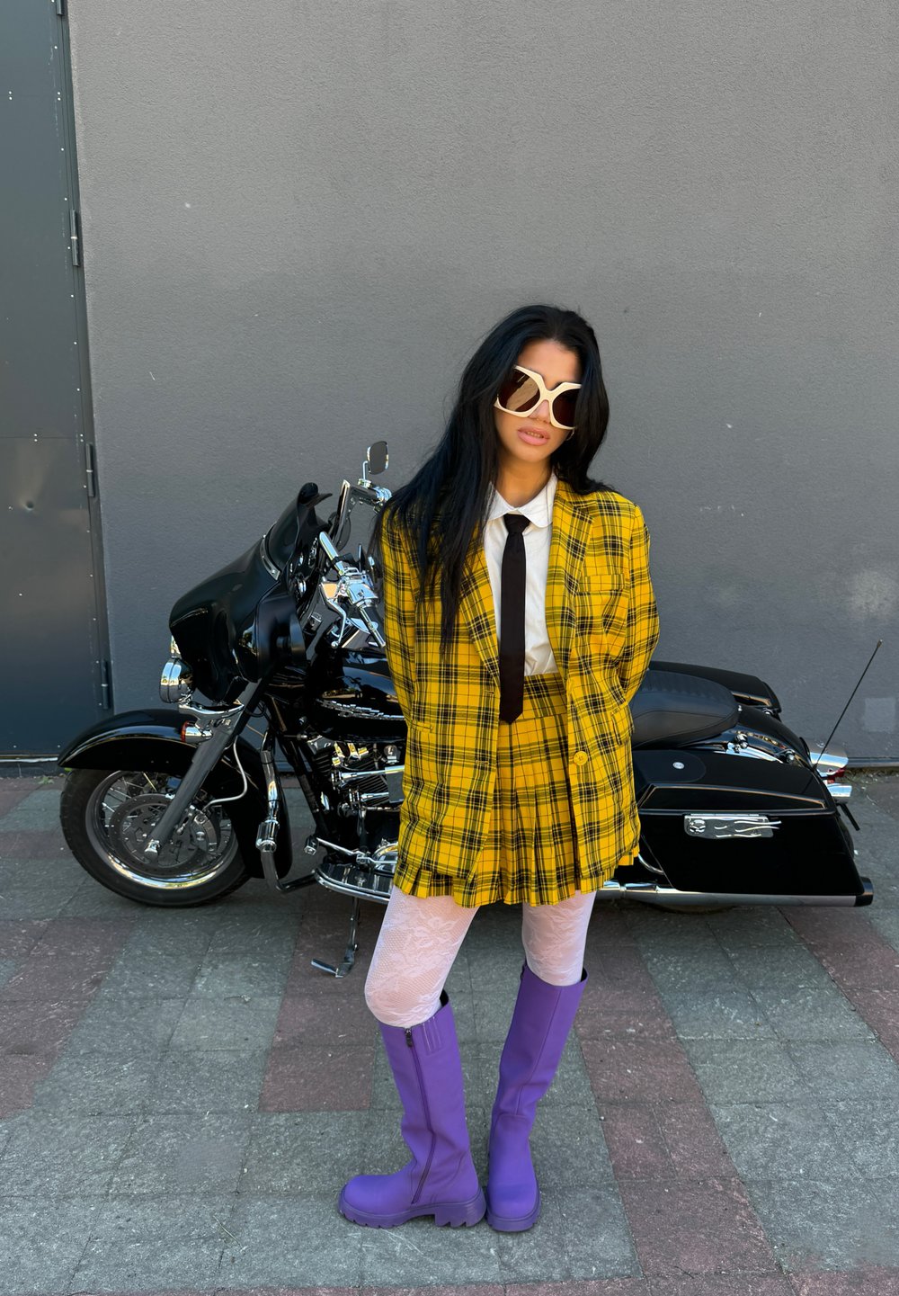 Blazer e saia xadrez amarelo, camisa branca com gravata preta, botas roxas até o joelho, de pé ao lado de uma motocicleta preta, fundo texturizado.