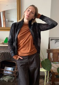 Črna puhasta bomberska jakna z visokim ovratnikom, nošena čez dolg rokav rjave barve in ohlapne črne hlače. Naravno notranje okolje.