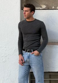 Νεαρός άνδρας με κοντά σκούρα μαλλιά, φορώντας εφαρμοστό σκούρο γκρι μακρυμάνικο πουκάμισο και ανοιχτό μπλε τζιν, στέκεται μπροστά σε έναν υφασμάτινο λευκό τοίχο.