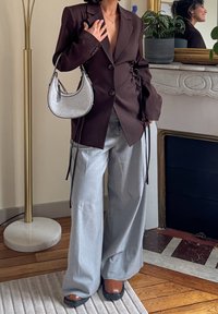 Blazer marron ajusté avec des détails froncés sur les côtés, associé à un pantalon large gris clair et un sac à main argenté métallique. Chaussures marron à talons épais.