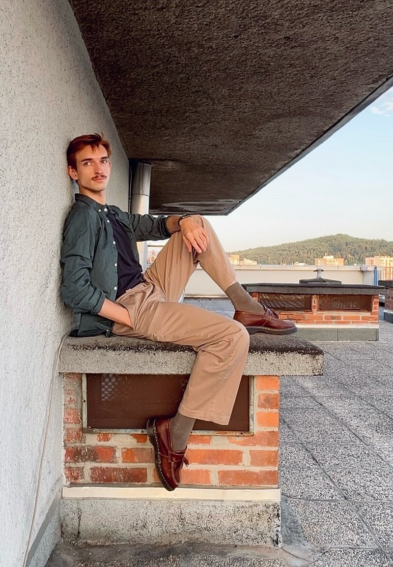 Jeune homme avec moustache assis sur le bord d’un toit, portant une chemise verte, un t-shirt noir, un pantalon beige et des chaussures marron, avec des collines en arrière-plan.