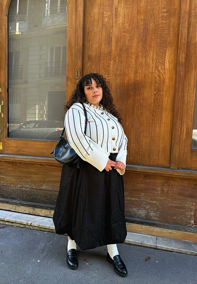 Cárdigan corto de rayas blancas con botones de madera, combinado con una falda larga negra. Bolsa de hombro negra y mocasines negros, de pie sobre la acera.
