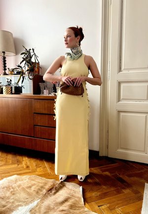 Vestido amarillo sin mangas con lazos laterales, combinado con un bolso marrón y una bufanda verde claro. Zapatos de tacón blancos completan el atuendo.