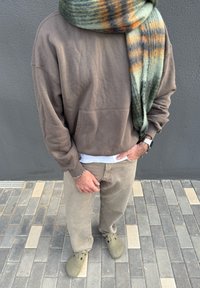 Sweat-shirt en polaire marron, pantalon gris, sabots vert olive et une écharpe à motifs verts, orange et marron drapée autour du cou.