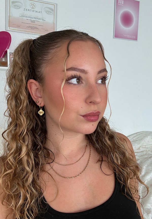 Jeune femme aux cheveux bouclés attachés en arrière, portant des boucles d'oreilles et des colliers en or, regardant sur le côté contre un mur blanc avec des certificats.