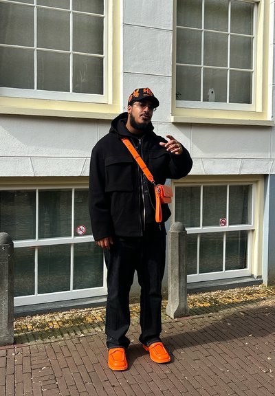 Chaqueta negra de gran tamaño, pantalones negros y zapatillas naranja brillante. Los accesorios incluyen un bolso cruzado naranja y una gorra a juego con logotipos.