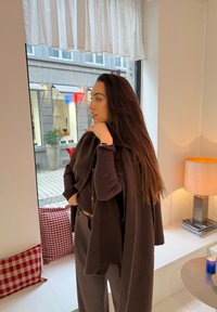 Cardigâ de malha castanho sobre uma blusa escura, combinado com calças cinzentas. Características notáveis incluem mangas longas e um corte casual, com iluminação interior quente.