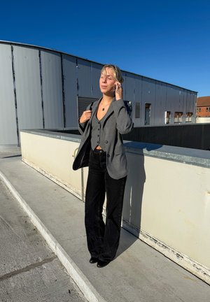 Blazer gris sobre una camisa de botones gris, pantalones de campana negros y zapatos negros. El entorno es al aire libre con un cielo azul claro y edificios.