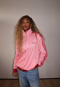 Jaqueta de treino rosa da Adidas com gola alta, fecho em zíper e detalhes em vermelho; combinada com jeans azul claro. Tecido suave com design esportivo.