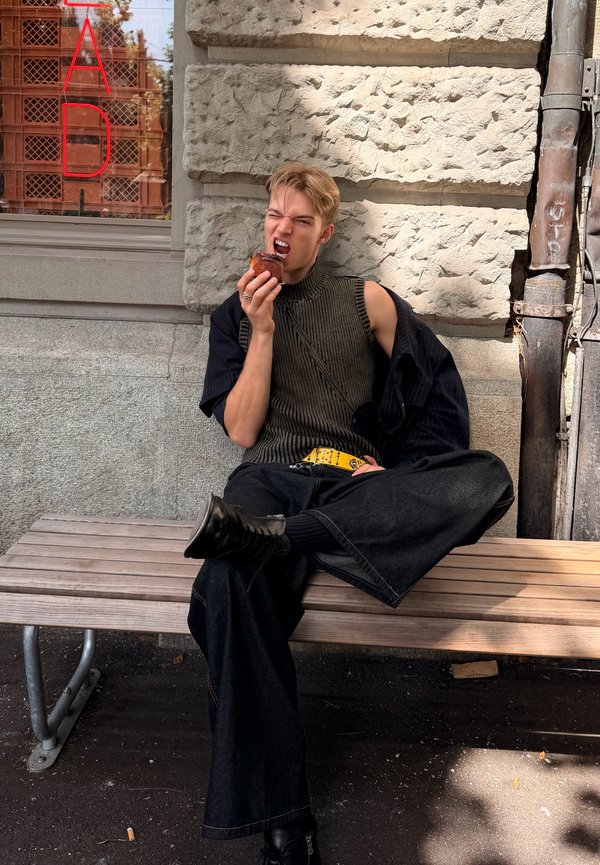 Jeune personne en pull sans manches et pantalon large assise sur un banc en plein air, mordant dans une pâtisserie.