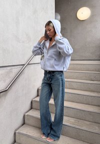 Harmaa vetoketjuhuppari, jonka sisäpuoli on valkoinen, yhdistettynä löysiin sinisiin farkkuihin. Malli seisoo portailla, pitäen hupparia kädessään, minimalistisessa taustassa.