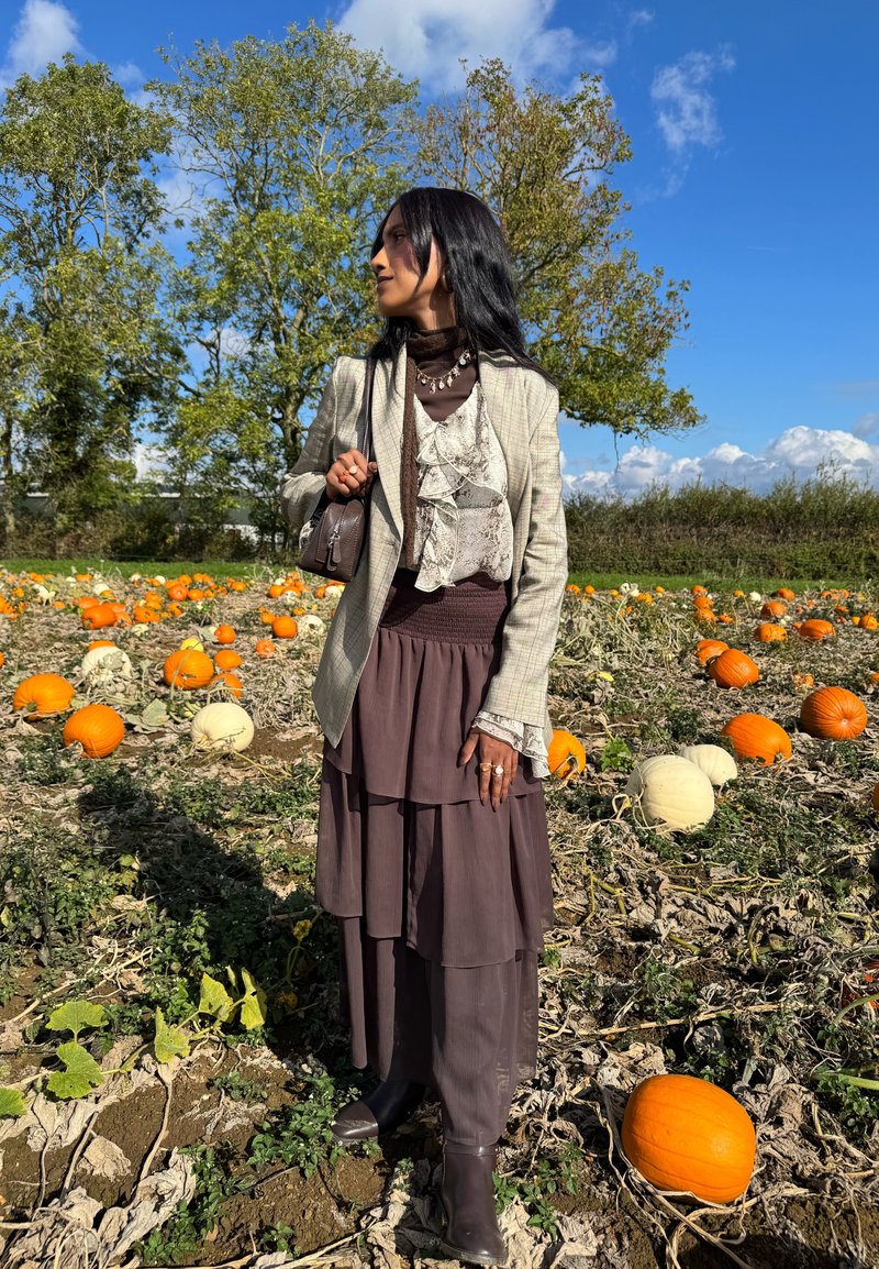 A layered brown skirt and top with ruffles, matched with a grey checked blazer, accessorised with a small bag and jewellery in a pumpkin field.