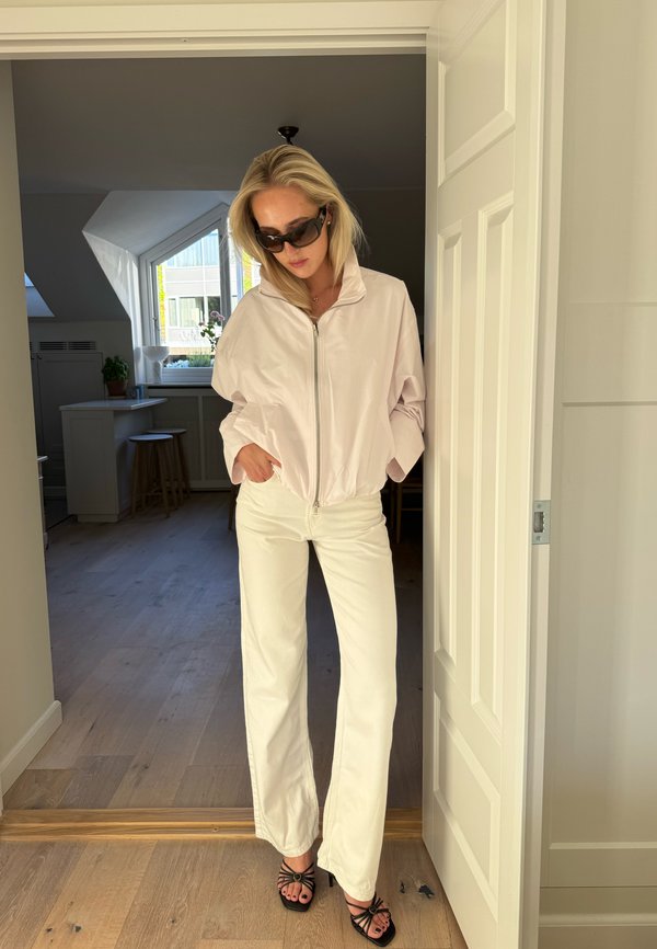 Femme portant une veste légère, un pantalon blanc et des sandales noires, debout dans une porte, les mains dans les poches, regardant vers le bas avec des lunettes de soleil foncées.