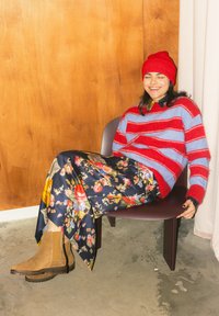 Personne souriante portant un pull rayé rouge et bleu, un bonnet rouge, une jupe fleurie et des bottes marron, assise confortablement sur une chaise contre un mur en bois et un rideau.