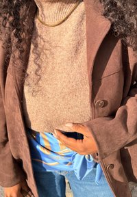 Personne aux cheveux bouclés portant un manteau en daim marron, un pull col roulé beige, un collier chaîne en or, une écharpe bleue à motifs nouée à la taille, et un jean bleu.