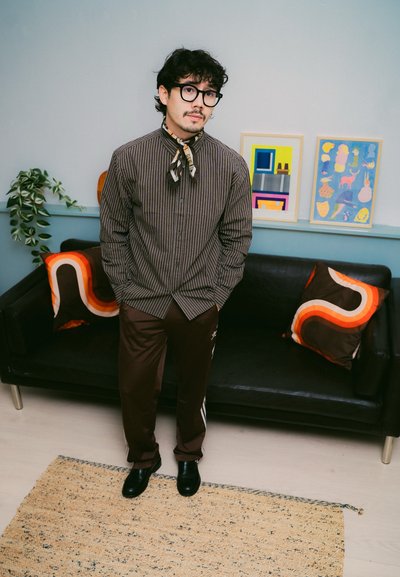 Camisa de botones a rayas, pantalones deportivos marrones con rayas laterales, calzado negro, bufanda estampada y un fondo de arte abstracto y cojines.