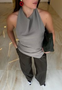 Hellgrau haltertop mit drapiertem Design, kombiniert mit dunkelgrünen weiten Hosen, hält eine strukturierte grüne Clutch und trägt rote, runde Ohrringe.