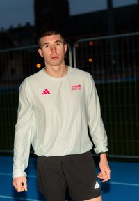 Jeune homme portant un t-shirt BMW Berlin Marathon à manches longues blanc et un short noir, debout sur une piste extérieure bleue au crépuscule.