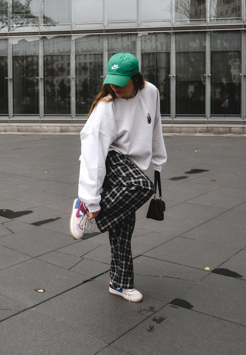 Λευκό oversized φούτερ με λογότυπο, παντελόνι καρό σε μαύρο και άσπρο, και πολυχρωματικά αθλητικά παπούτσια. Συμπεριλαμβάνεται ένα πράσινο καπέλο και μία μικρή μαύρη τσάντα.