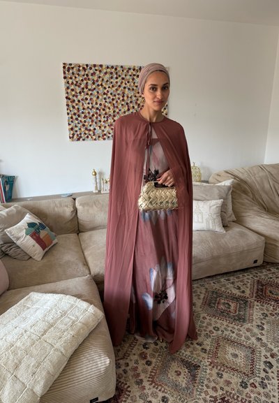 Mujer con pañuelo malva en la cabeza y capa sobre un vestido floral, sosteniendo un bolso dorado tejido, de pie en una sala de estar beige con alfombra estampada.