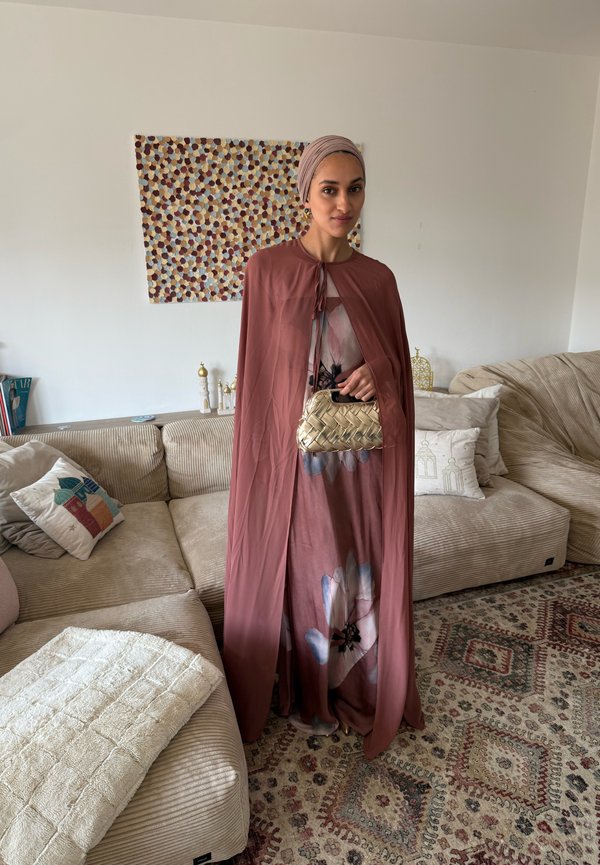 Kobieta w mauve chustce na głowie i płaszczu narzuconym na kwiecistą sukienkę, trzymająca plecioną złotą kopertówkę, stojąca w beżowym salonie z dywanem w wzory.