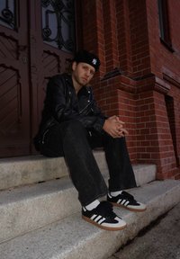 Zwarte leren motorjack, zwarte spijkerbroek en zwart-witte sneakers met rubberen zolen, zittend op stenen traptreden tegen een achtergrond van rode bakstenen.