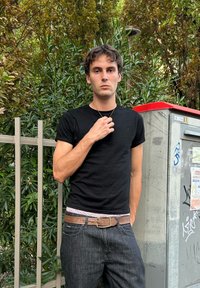 Black short-sleeve t-shirt, dark denim jeans, brown belt with a silver buckle, standing near a green leafy background and a grey utility box.
