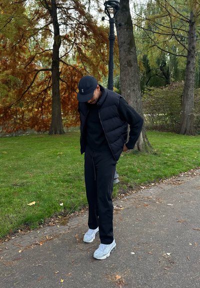 Chaleco negro acolchado sobre un suéter negro, combinado con pantalones negros y zapatillas gris claro, de pie en un sendero rodeado de árboles otoñales.