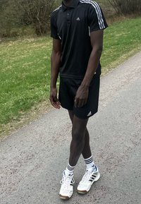 Fekete Adidas póló fehér csíkokkal, fekete rövidnadrág, fehér sportcipő kék részletekkel és csíkos zokni, egy füves úton elhelyezve.