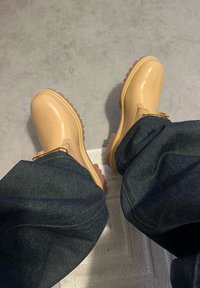 Beigenväriset vedenpitävät saappaat, joissa on kiiltävä viimeistely, paksu kuvioitu pohja, nauhat ja kultaiset reiät, yhdistetty tummansinisiin farkkuihin.