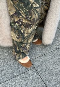 Κάτω πόδια με καμουφλάζ παντελόνια cargo και καφέ παντόφλες με επένδυση γούνας, που στέκονται σε γκρι πλακάκια δίπλα σε γούνινο παλτό ανοιχτού χρώματος.