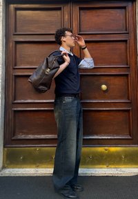 Borsa di pelle marrone con zip e manico, indossata sulla spalla. Outfit informale con una maglietta blu navy, camicia azzurra chiara e jeans blu scuro.