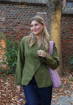 Blazer verde oliva con rayas finas y un solo botón, combinado con vaqueros oscuros. Accesorios con un bolso rosa y joyas doradas.