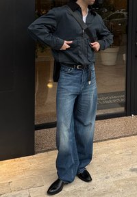 Personne portant une veste noire, un jean large bleu délavé, des bottes noires, ajustant la sangle d'un sac bandoulière noir à l'extérieur d'une entrée de bâtiment.