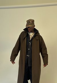 Mann in einem langen braunen Mantel über einer dunklen Jacke und Hose, Camouflage-Mütze, Sonnenbrille und mehreren goldenen Ketten, der vor einer schlichten Wand steht.