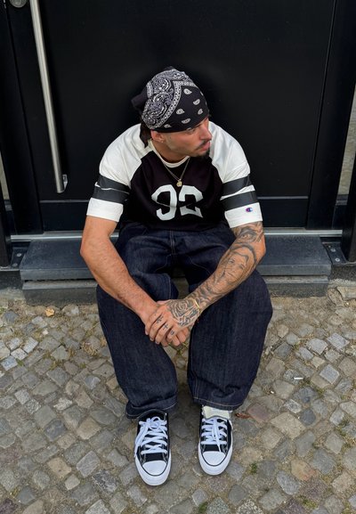 Camisa de manga corta en blanco y negro con el número 93 en negrita, jeans azul oscuro, zapatillas blancas y negras, y una bandana negra, sentado sobre adoquines.