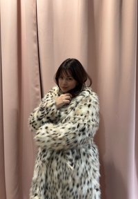 Jeune femme aux cheveux foncés portant un manteau en fausse fourrure blanc à taches noires, debout devant des rideaux rose pâle.