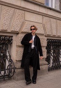 Melns virsdrēbes mētelis, svītraini krekls, balts T-krekls, melnas bikses un saulesbrilles. Gludas tekstūras; ievērojams zīmola josta. Fona akmens siena.