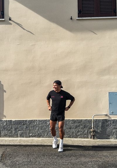 Hombre sonriente con ropa deportiva negra y una gorra hacia atrás está de pie con las manos en las caderas frente a una pared beige al aire libre en un día soleado.