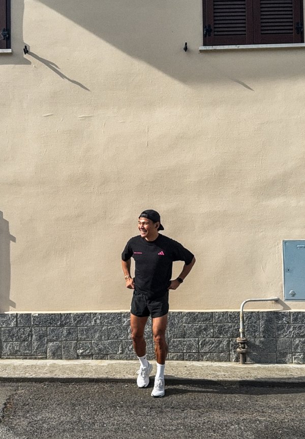 Nasmiješeni muškarac u crnoj sportskoj odjeći i s kapom unatrag stoji s rukama na bokovima ispred bež zida vani na sunčan dan.