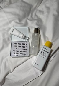 Vier Hautpflegeprodukte auf weißem Stoff angeordnet: eine silberne Dose, eine weiße Tube, eine klare Flasche mit silbernem Deckel und eine weiße Flasche mit gelbem Deckel.