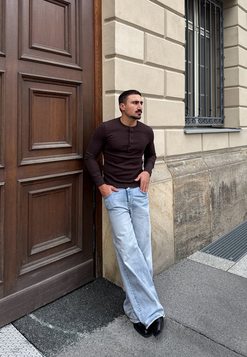 Homme aux cheveux foncés et à la barbe, portant une chemise noire à manches longues et un jean bleu clair, appuyé contre un mur en pierre beige proche d'une grande porte en bois.