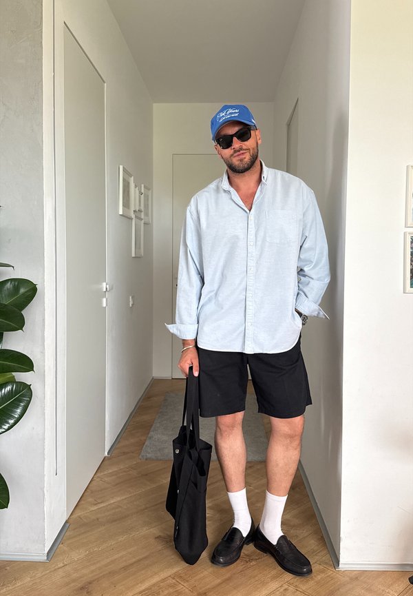 Mand iført blå kasket, solbriller, lyseblå skjorte, sorte shorts, hvide sokker og sorte sko, står i gangen og holder en sort mulepose.