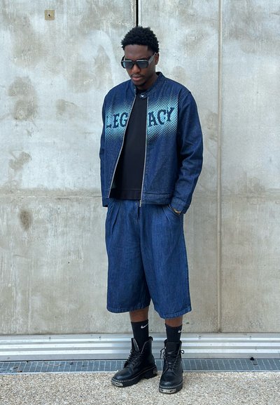 Chaqueta de mezclilla con letras en degradado azul, camiseta negra, pantalones cortos de mezclilla holgados y botas de combate negras. Corte moderno y estilo casual.