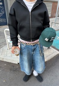 Must käibemüts, valge T-särk, ruuduline ja leopardimustriga vöö, sinised teksashortsid, mustad sussid, rohelise mütsi all, käes jääjoogiga.