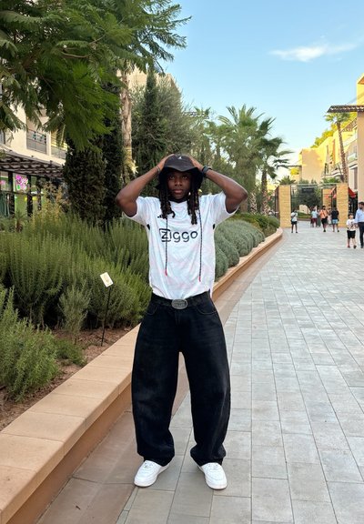 Camisa blanca de manga corta con un colorido patrón de olas y la impresión "Ziggo"; jeans negros de pierna ancha; sombrero de cubo negro; zapatillas blancas.