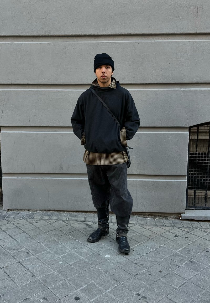 Black oversized jumper with a checkered shirt underneath, dark trousers, and black boots. The person stands against a grey wall.