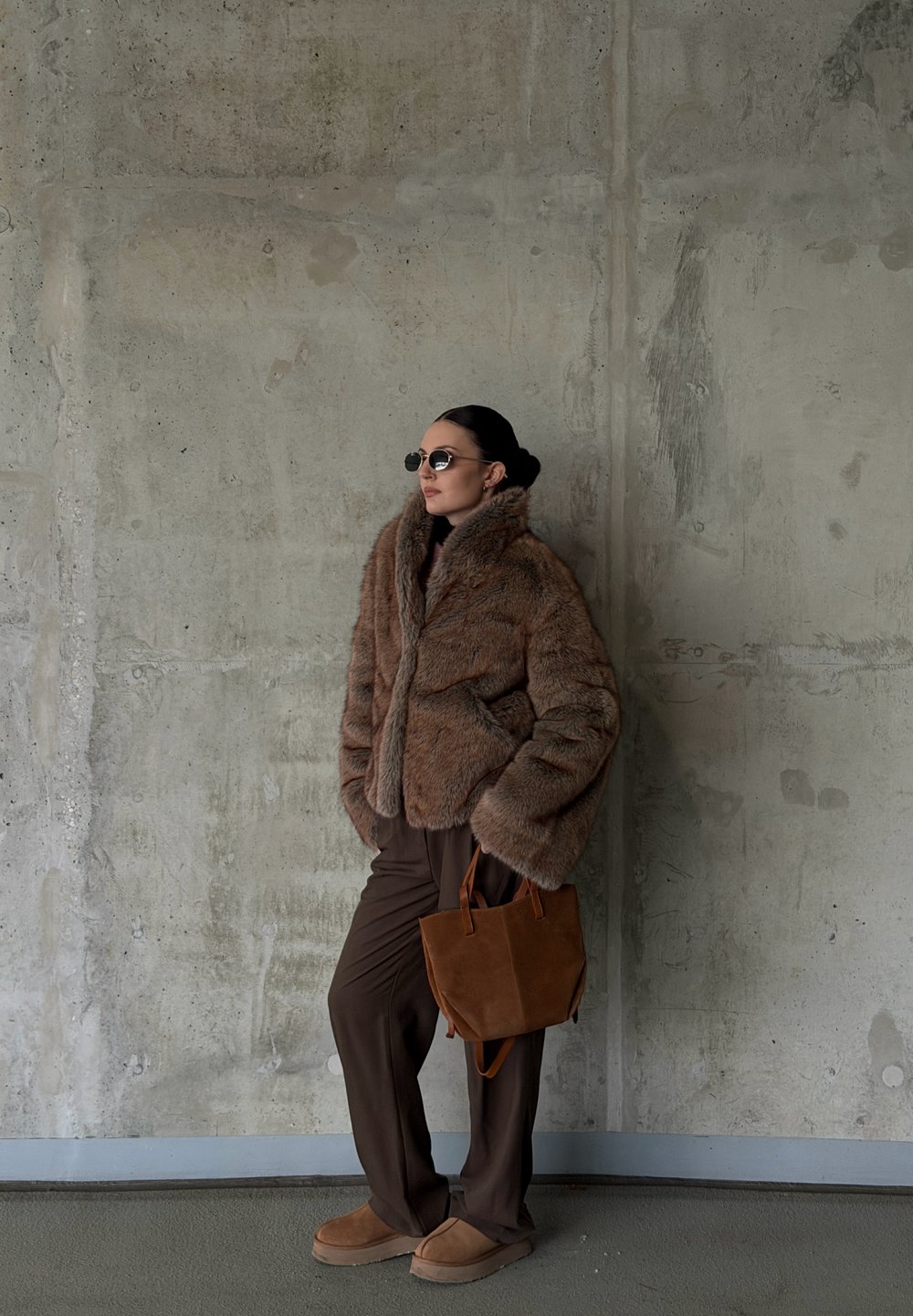 Veste en fausse fourrure marron, pantalon marron foncé, chaussures en daim marron clair et petit sac à main marron ; des lunettes de soleil rondes complètent le look sur un fond en béton.
