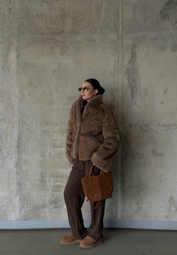 Hnedá bunda z umelého kožušinového materiálu, tmavohnedé nohavice, topánky z hnedej semišovej kože a malá hnedá kabelka; okrúhle slnečné okuliare dotvárajú vzhľad na betónovom pozadí.