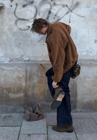 Brun ruskindsjakke, mørke denimjeans, grøn camouflage taske på jorden, person holder en brun ankelstøvle mens de justerer fodtøjet.